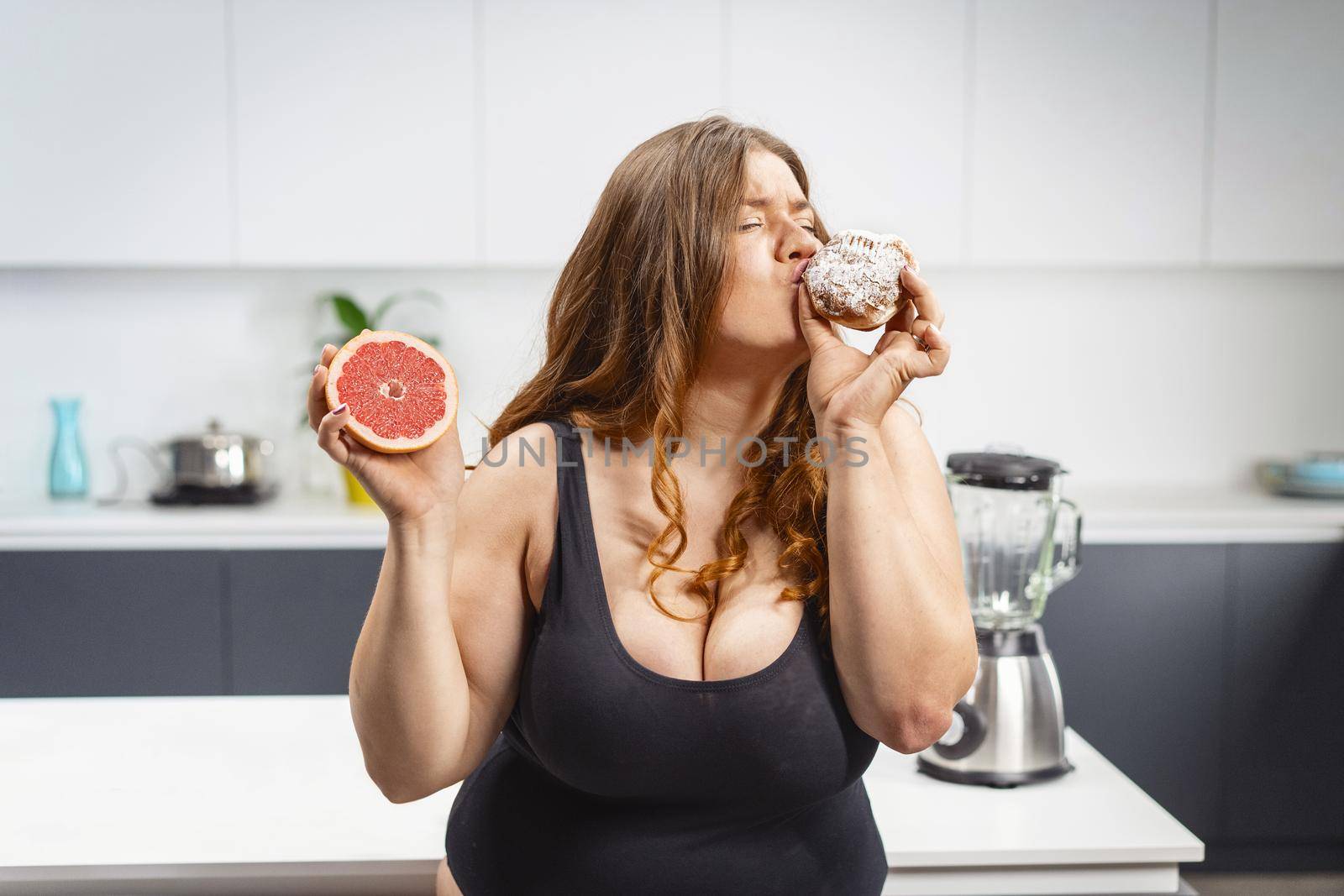 Young fat woman choosing what to eat holding a cake and grapefruit. Beautiful chubby young woman eating unhealthy food. Fat girl comparing between eating cake and fruit by LipikStockMedia