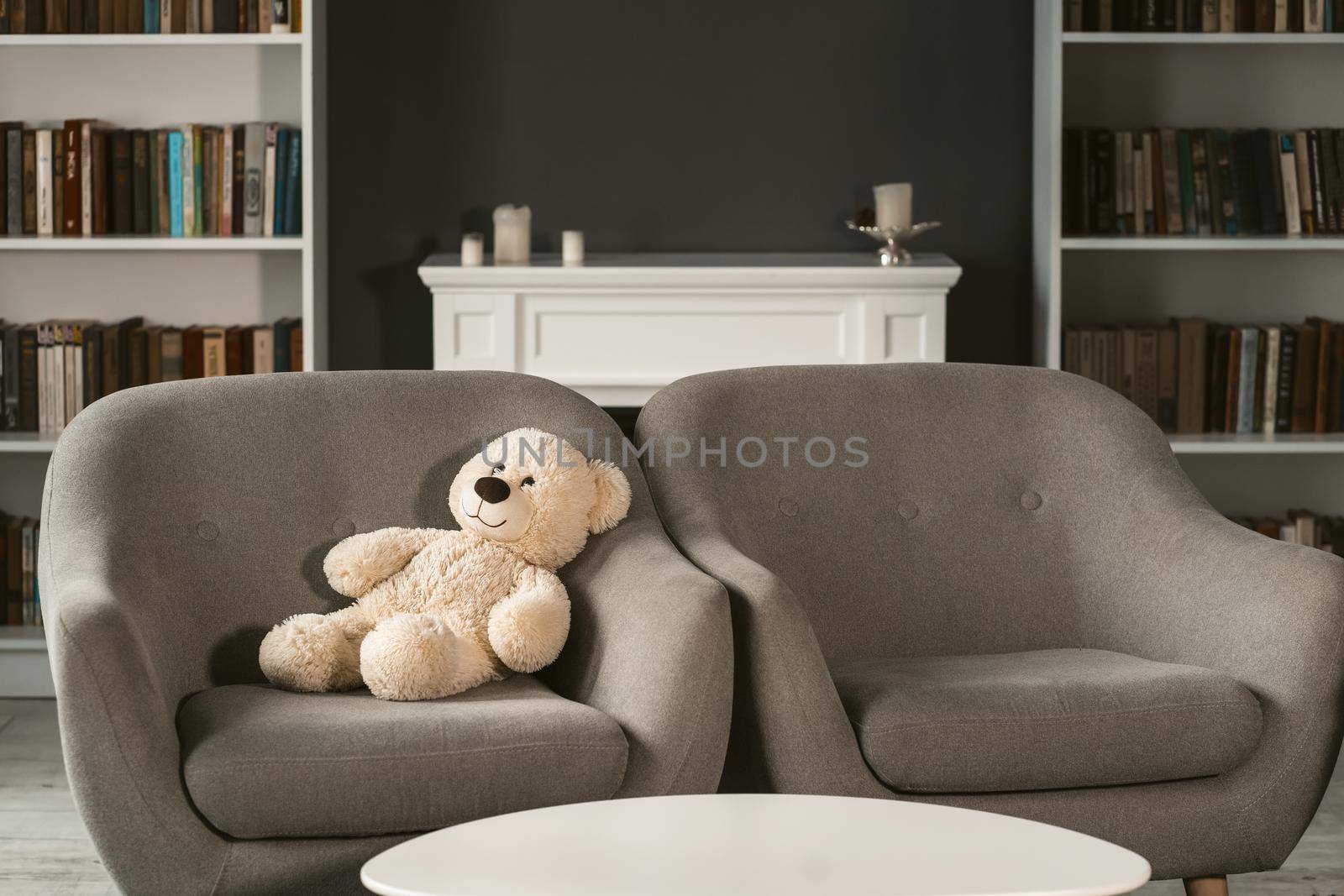 Teddy sitting on armchair. Teddy bear on grey chair next to empty one in interior. Beautiful living room with teddy bear on chair.