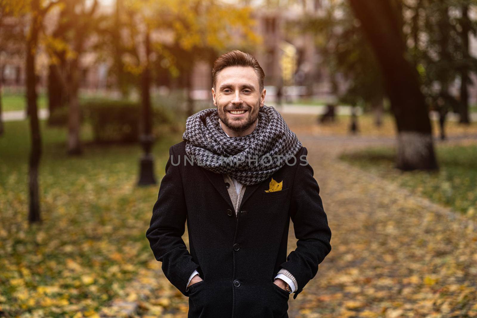 Smiling handsome young man looking at camera standing outdoors with hands in pockets of his dark blue coat. Young freelancer man stands on the in an autumn coat looking happy at camera.