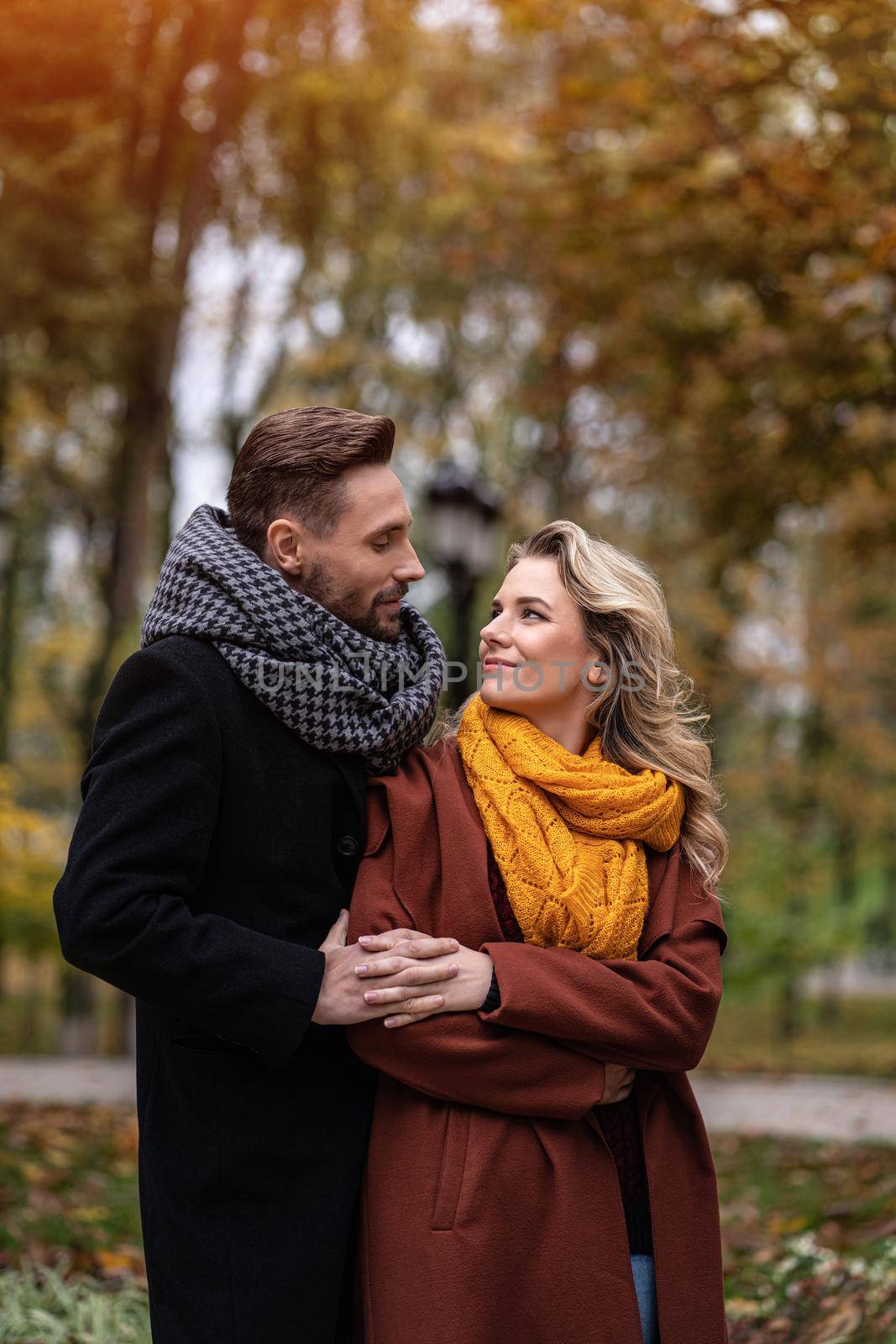 Handsome man and a woman hugged from behind smile looking at each other in the autumn park. Outdoor shot of a young couple in love having great time. Autumn toned image.