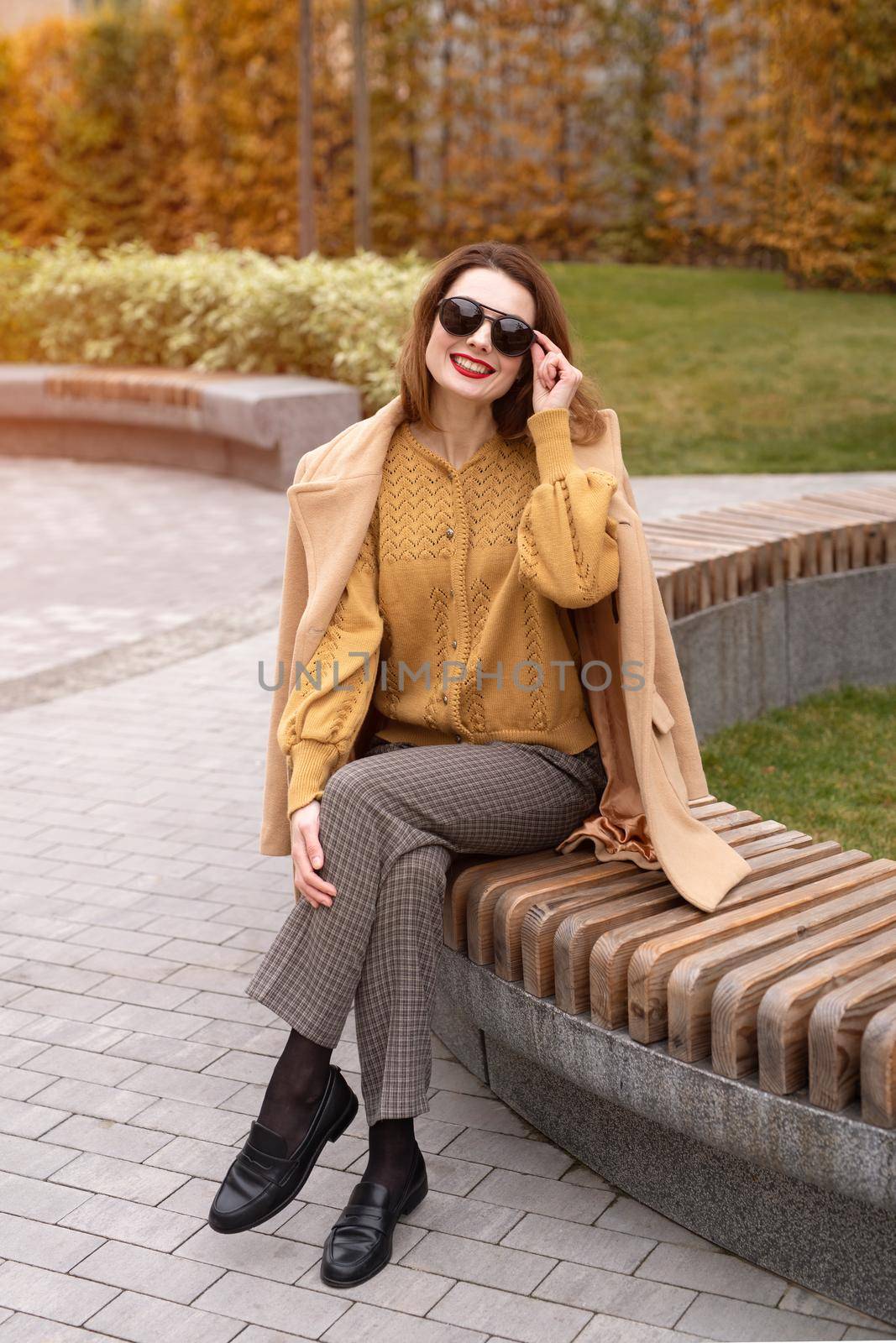 Beautiful young woman in an autumn beige coat and sunglasses sits on a flowerbed bench waiting for her date or girlfriends. Toned photo. 