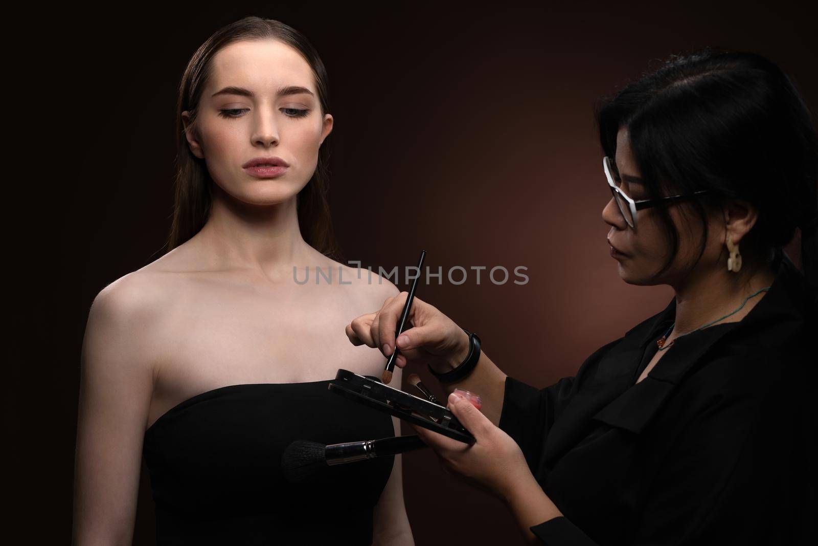 Beauty makeup artist working on a fashion model pretty face isolated on black background. Beautiful model at make up artist. Make-up artist, applying eye shadow to the model by LipikStockMedia