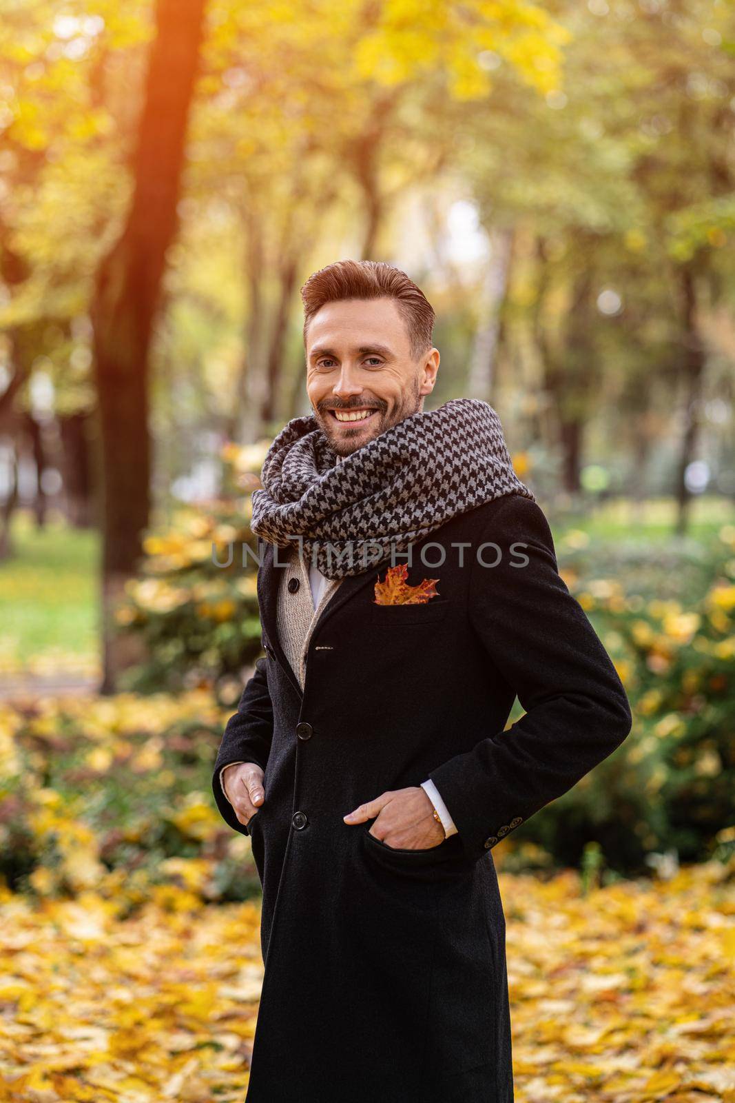 Handsome man wearing dark blue coat and scarf waiting on his date smile on camera. Young freelancer man stands on the street in an autumn coat looking at his woman.