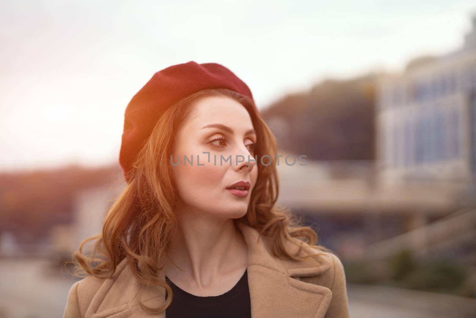 Charming pretty french woman in an autumn beige coat and red beret standing outdoors with urban city background. Tinted photo. 