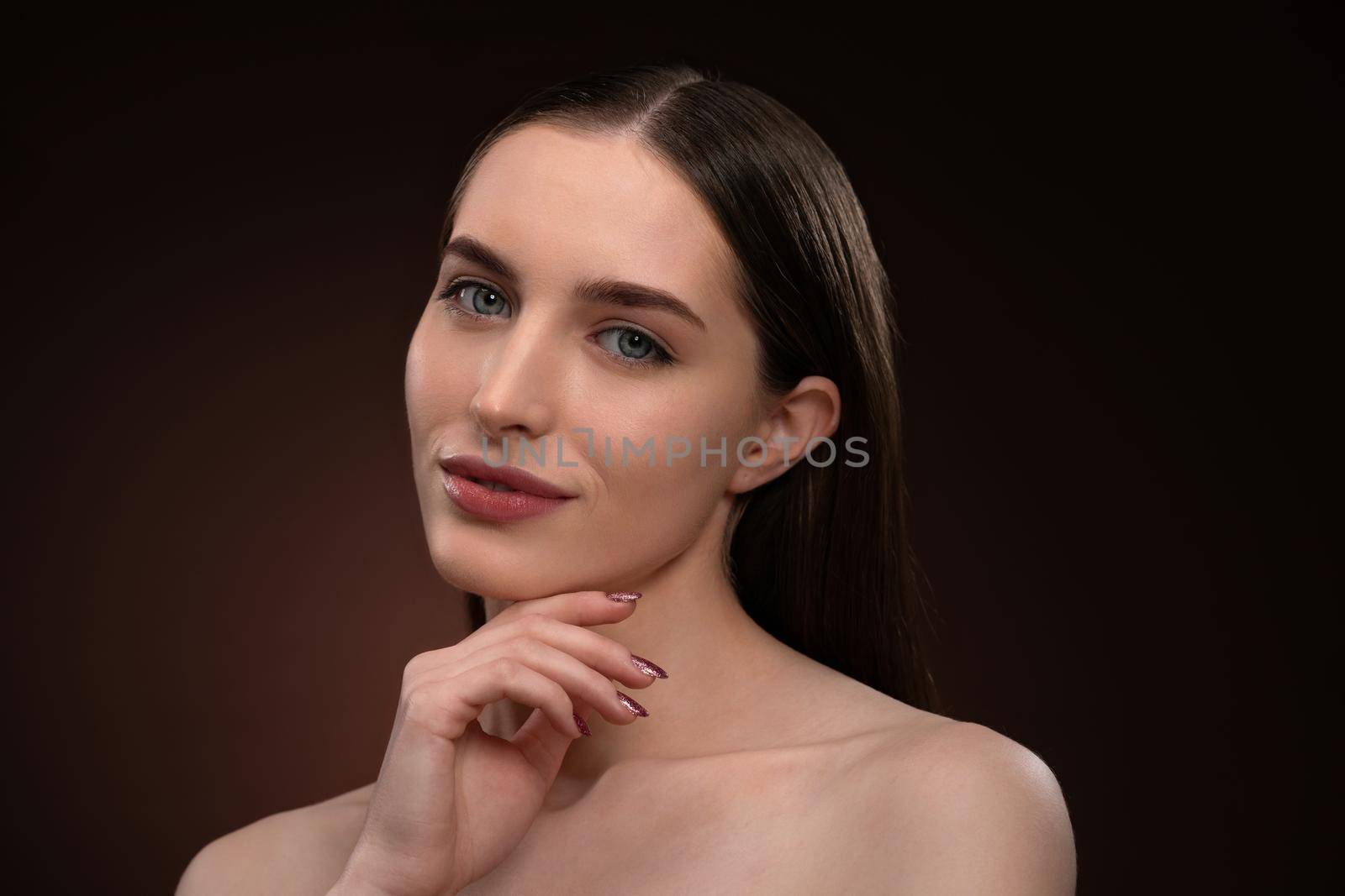 Beautiful spa model girl with perfect fresh clean skin and bare shoulders. Charming brunette model girl posing on camera touching her chin with a finger isolated on black background. 