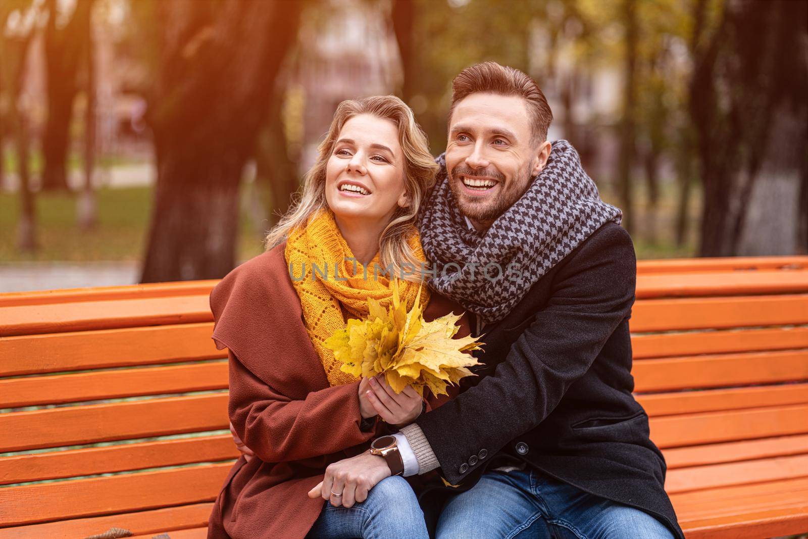 Charming embracing happy couple sitting on the bench romantic hugged in park wearing coats and scarfs Collecting a bouquet of fallen leaves. Love story concept. Tinted image. 
