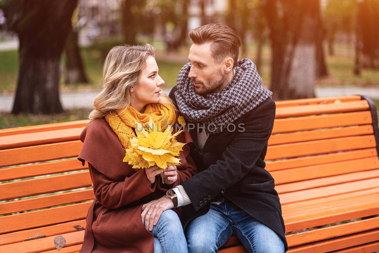happy loving couple embracing sitting on the bench romantic hugged in park wearing coats and scarfs Collecting a bouquet of fallen leaves. Love story concept. Tinted image by LipikStockMedia
