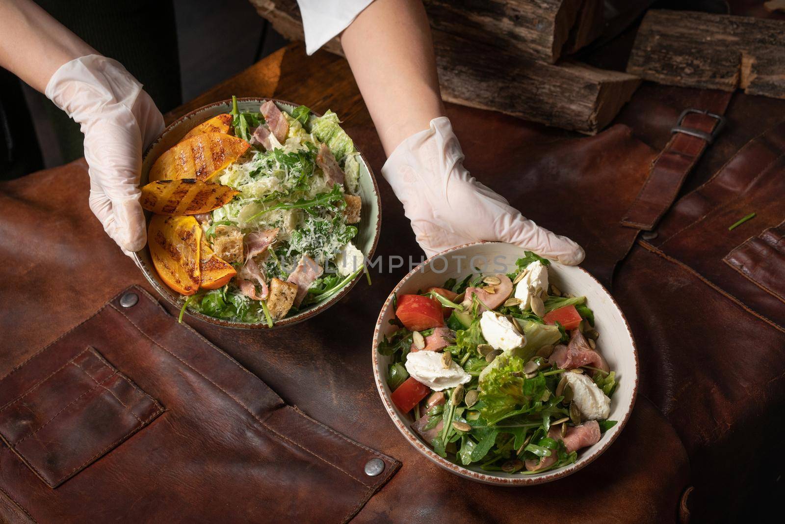 Two hands with bowls of vegetable salad with salmon, mozzarella cheese and grilled slices of fruits stands on a table covered with a leather rag. Restaurant concept. Vertical image by LipikStockMedia