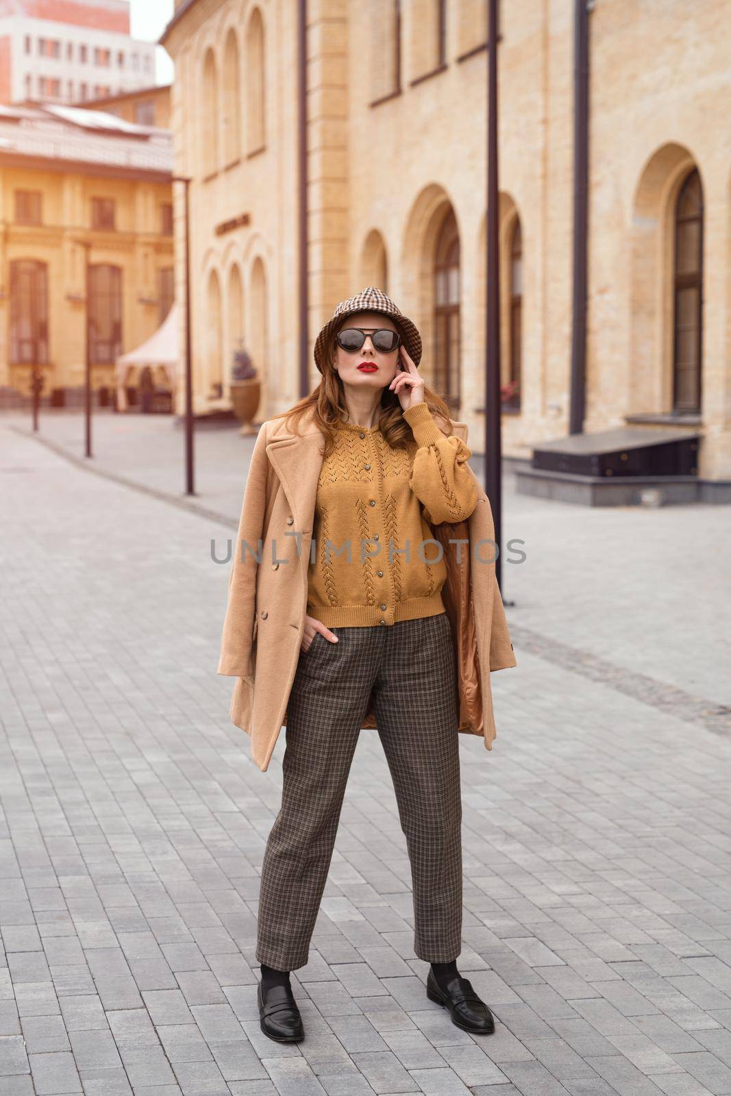 Full height self confident girl in an autumn beige coat and sunglasses, plaid panama hat standing posing on the street with coat on her shoulders. Retro toned photo. 