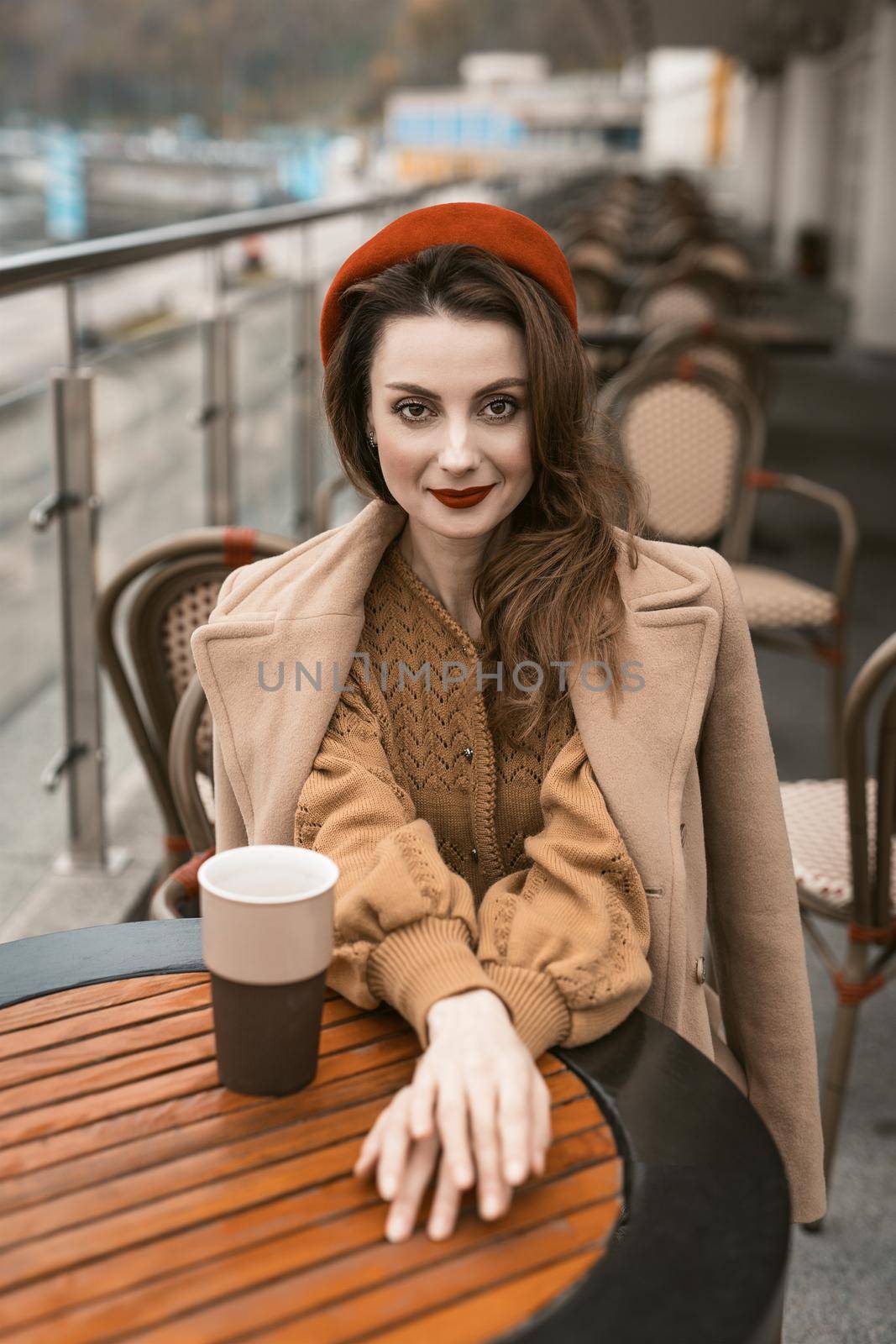 French woman drink coffee sitting at restaurant terrace with coffee mug looking at camera. Portrait of stylish young woman wearing autumn coat and red beret outdoors. 