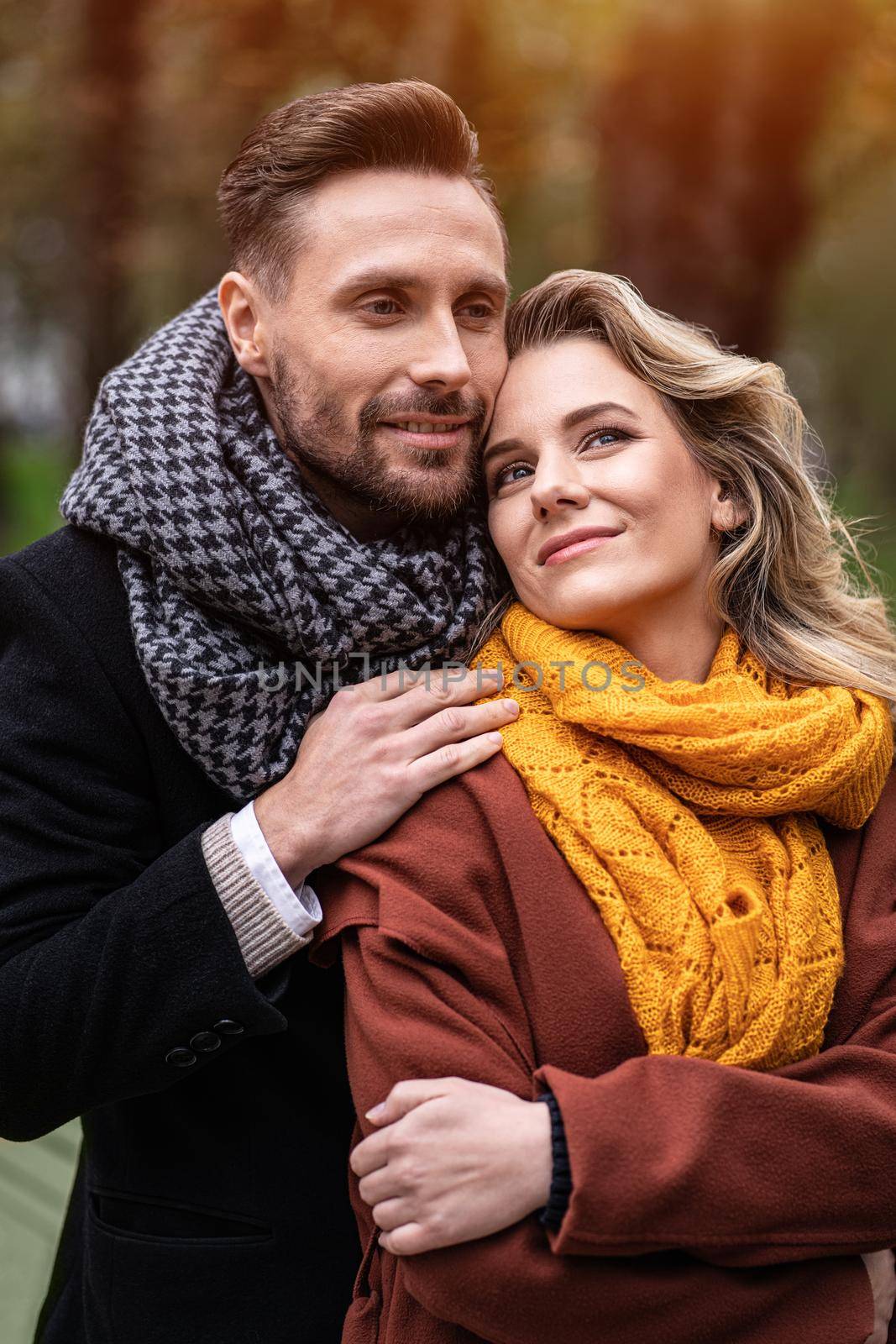 Tenderness of a cute couple. Handsome man and a woman hugged from behind smile looking at each other in the autumn park. Outdoor shot of a young couple in love having great time.