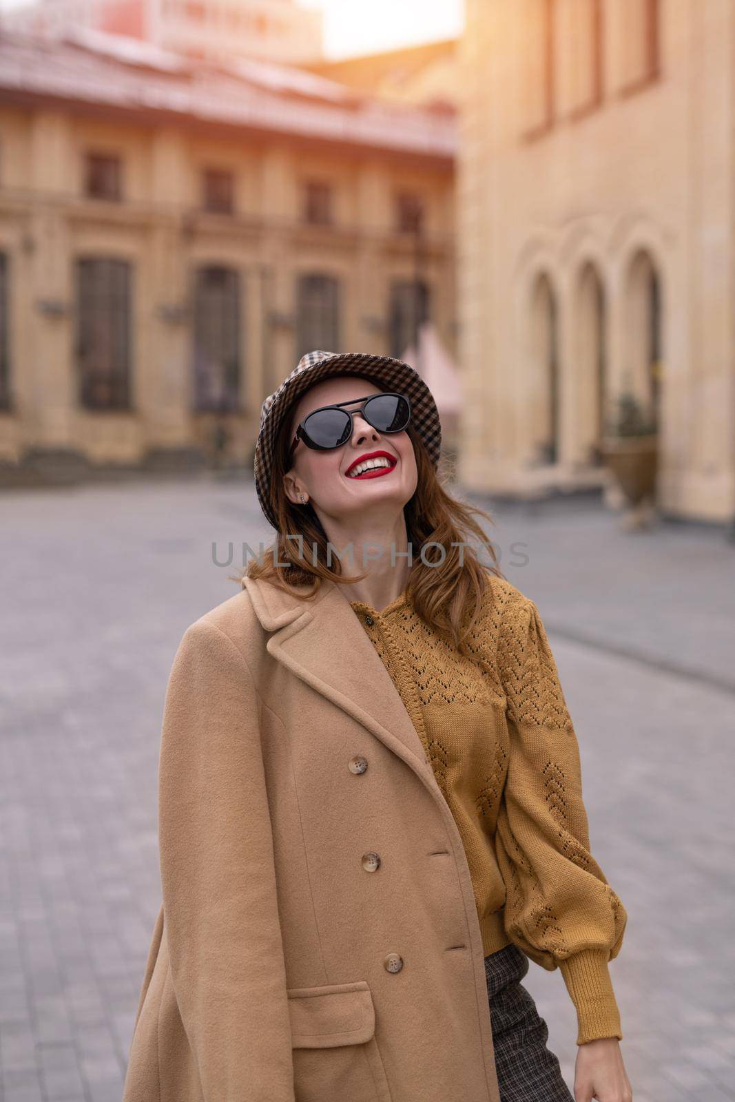 Charming girl in an autumn beige coat and sunglasses walking on the street happily posing for the camera. Vertical photo. Tinted photo by LipikStockMedia