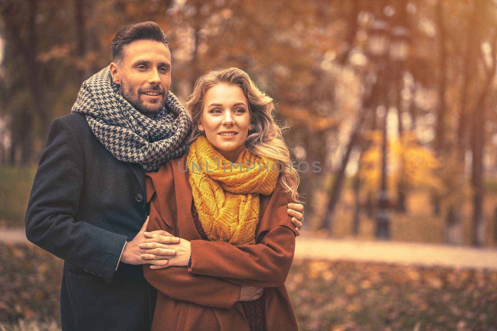 Couple in love man and a woman hugged from behind smile looking at their kids in the autumn park. Outdoor shot of a young couple in love having great time. Autumn toned image by LipikStockMedia