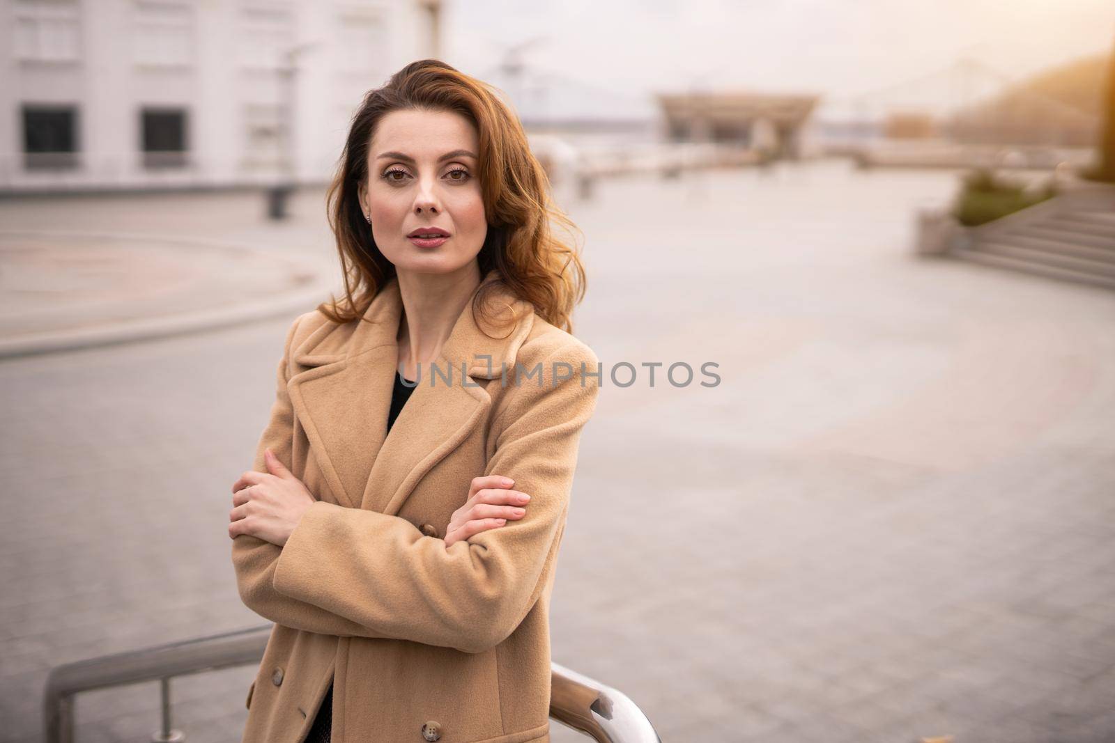 Charming young woman in an autumn beige coat standing leaned with her arms folded with urban city background. Toned photo. 