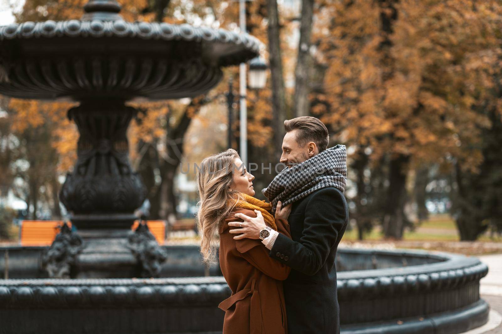 Beautiful couple of young people stands embracing and looking at each other near a beautiful fountain in the autumn park wearing autumn coats. Tinted image. 