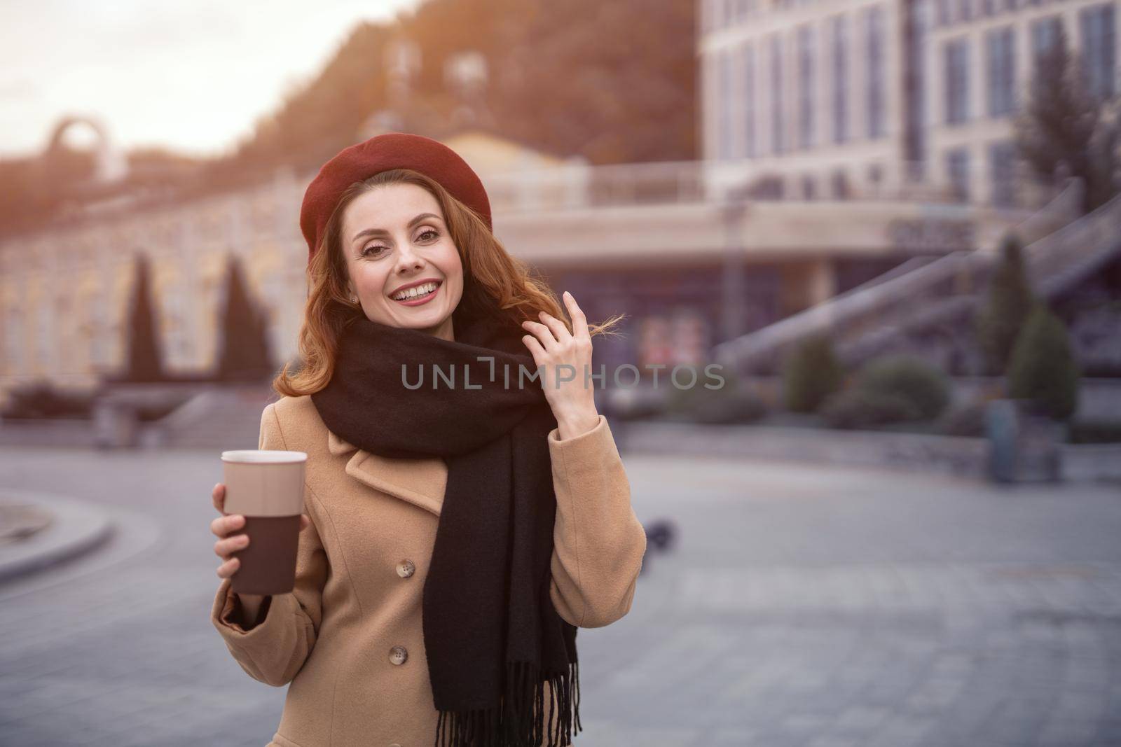 Casual portrait of french woman holding coffee mug outside in autumn beige coat and red beret with urban city background. Tinted photo by LipikStockMedia