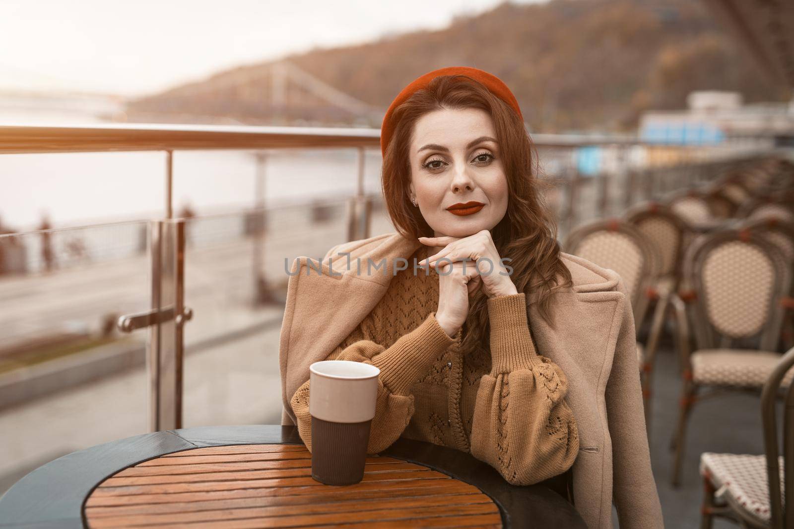 Lovely french young woman sitting at restaurant terrace with coffee mug looking at camera. Portrait of stylish young woman wearing autumn coat and red beret outdoors. 