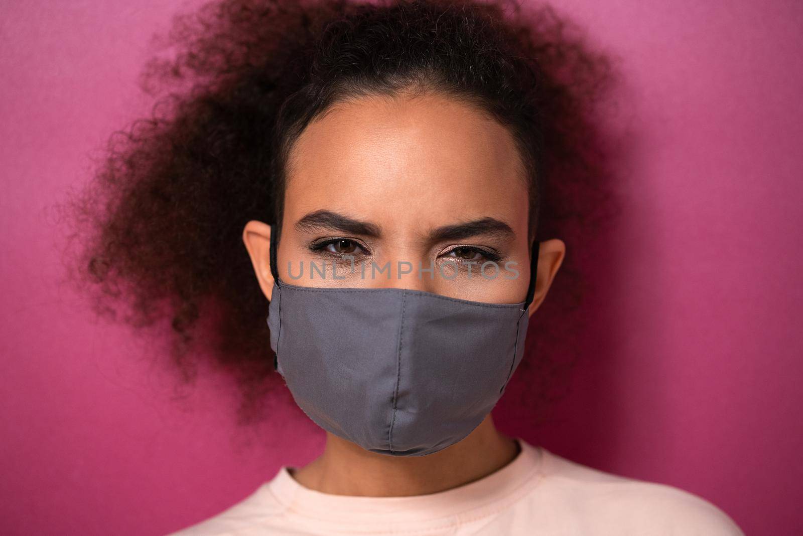 Questioning or studying look wearing reusable face mask African American charming girl in peachy t-shirt, to prevent others from corona COVID-19 and SARS cov 2 infection isolated on pink background by LipikStockMedia