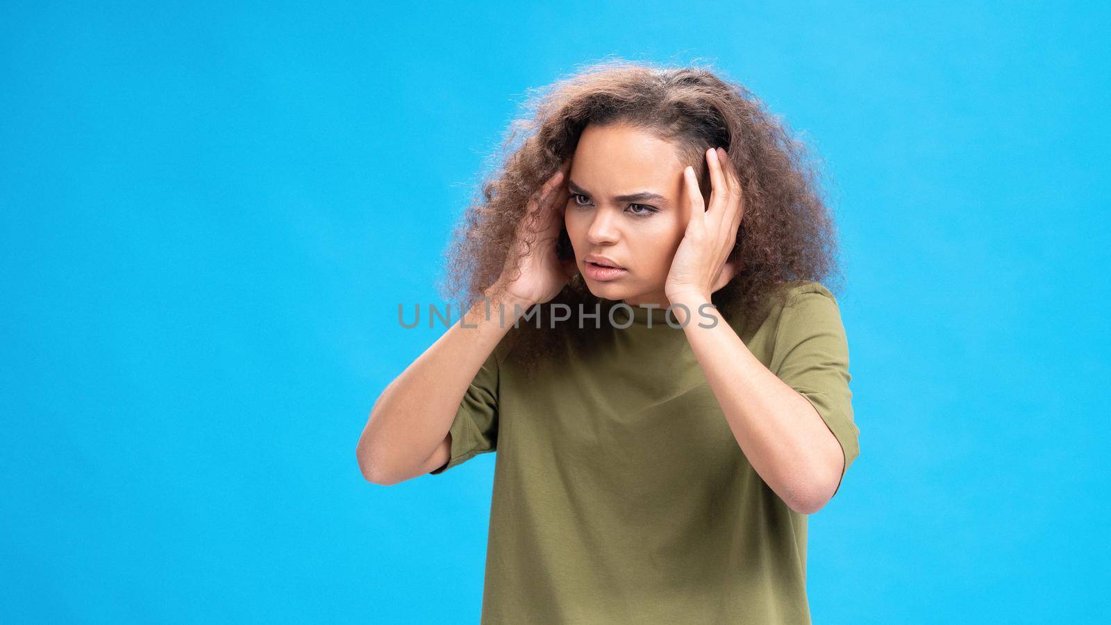 Stressed or unhappy african american girl touching her head suffering from headache wearing olive t-shirt isolated on blue background. Beauty concept. Health care concept. 