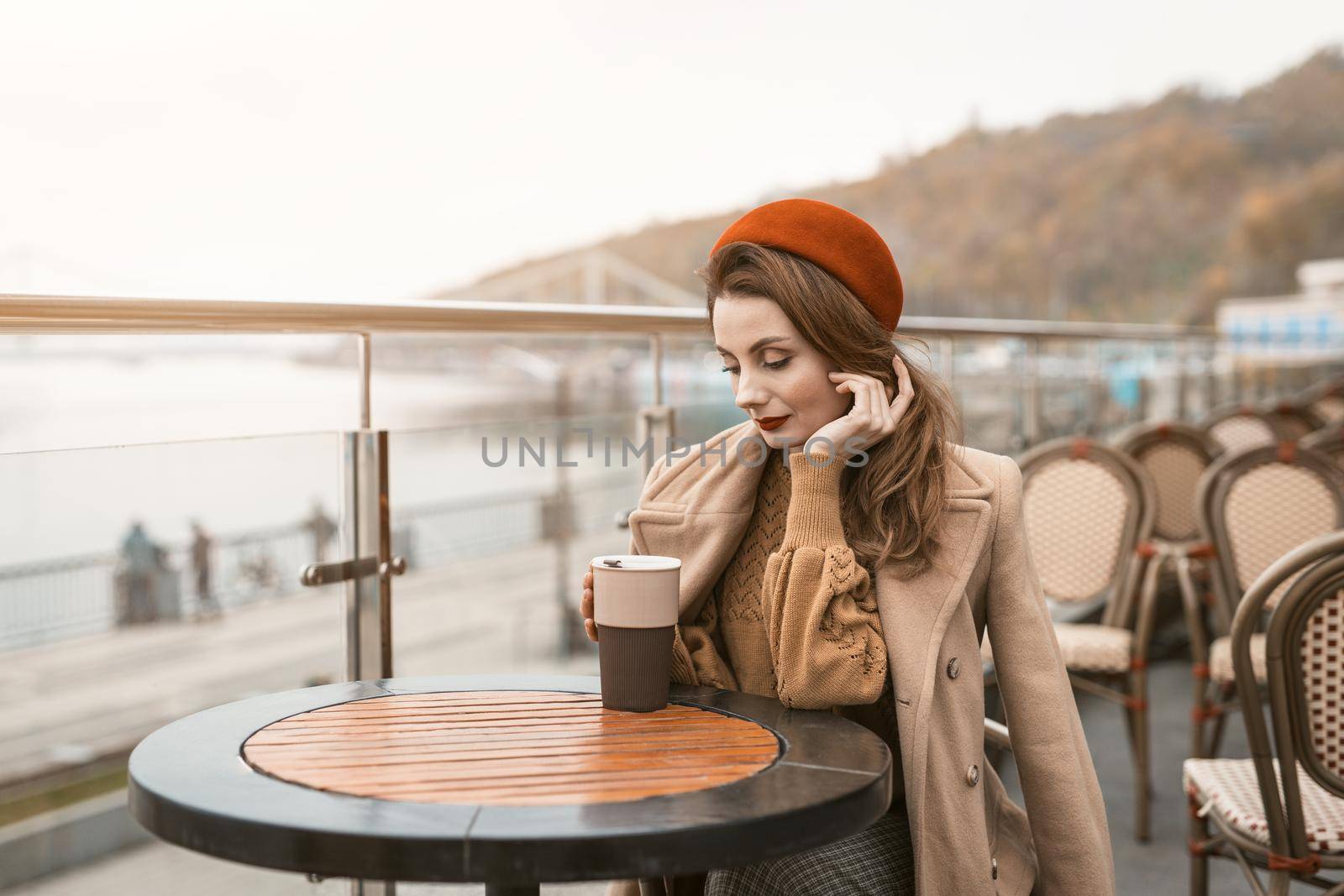 Thoughtful young woman looking at cup of coffee outdoors. Frenchwoman wearing red beret sitting on terrace of restaurant or cafe. Autumn urban city on background. 