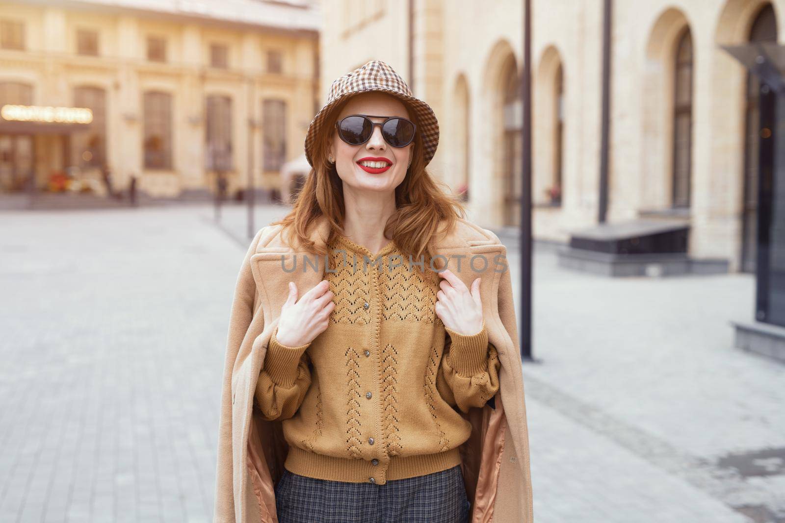 Charming girl in an autumn beige coat and sunglasses walking on the street with coat on her shoulders. Toned photo. 