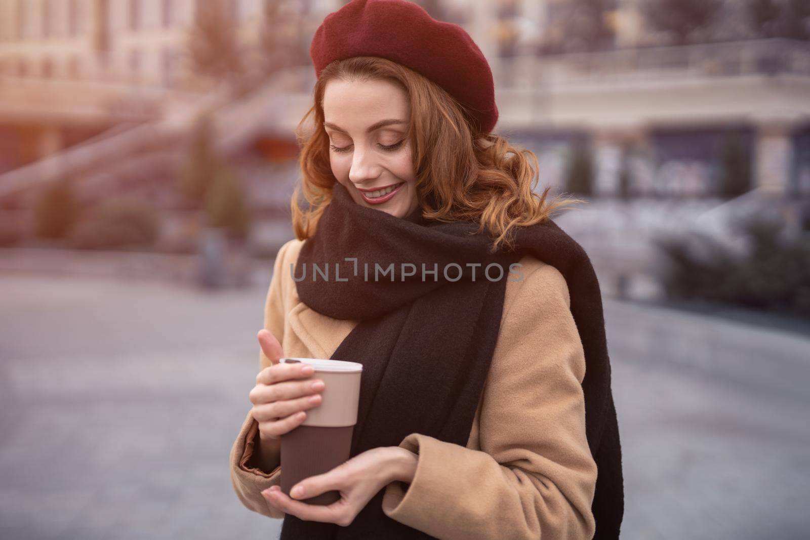 Half-length portrait of a beautiful french styled young woman holding a coffee mug standing outdoors. Happy french stylish young woman wearing autumn coat and red beret. Tinted image by LipikStockMedia