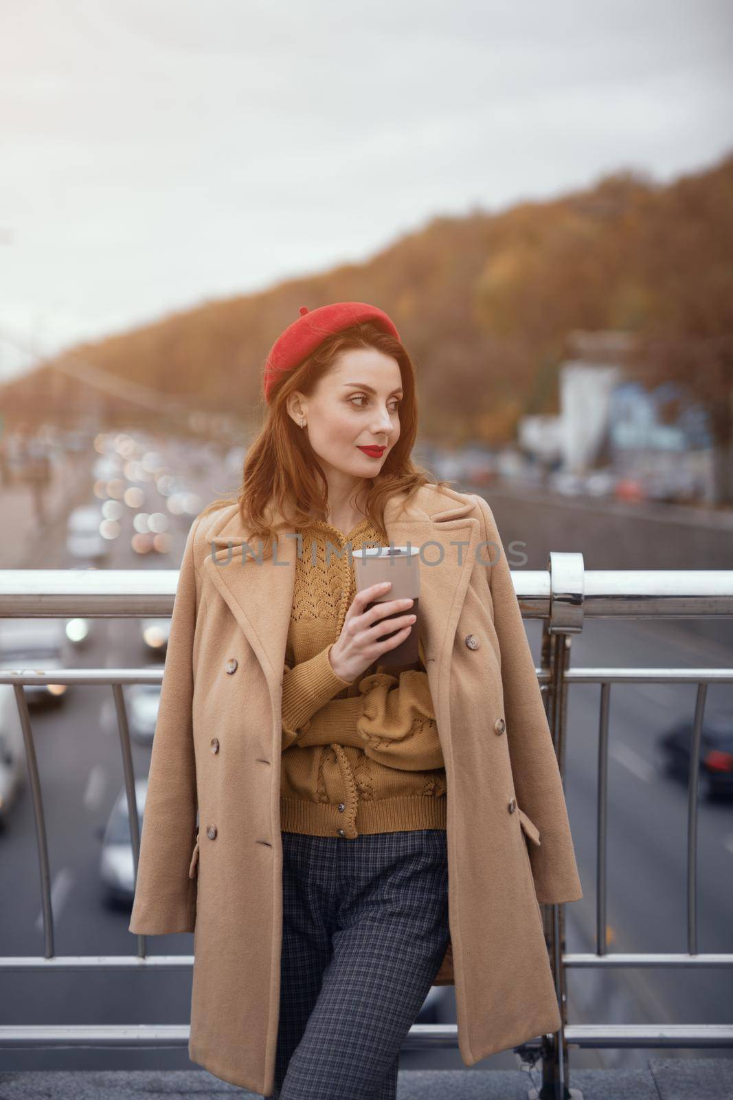 Tender young woman warming up with cup of coffee on the street female fashion standing on a pedestrian bridge. Portrait of stylish young woman wearing autumn coat and red beret outdoors. Toned image by LipikStockMedia