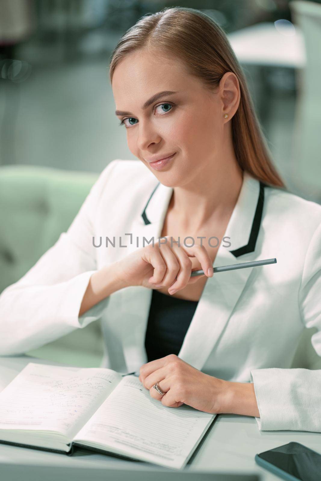 Beautiful businesswoman in white jacket looking at camera making notes in her scheduler or journal sitting in front at the modern bright office. Office worker looking at her working place by LipikStockMedia