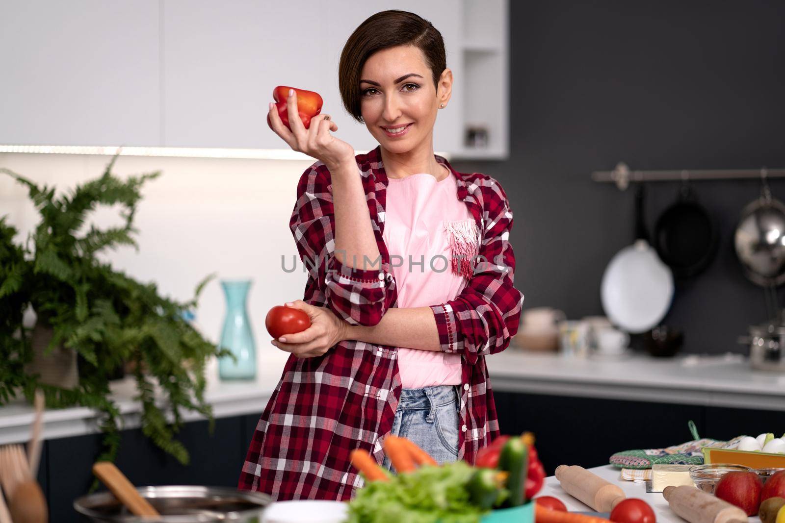 Holding bell pepper and tomato in the hand housewife, wearing plaid shirt with a short hairstyle cooking vegetable ragout at new kitchen in new house. Healthy food at home. 