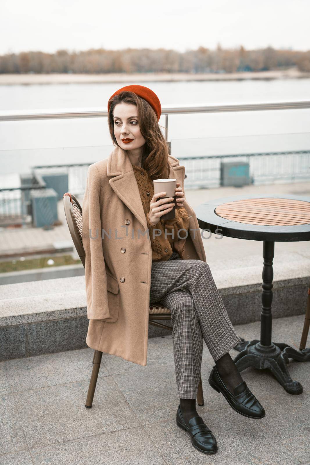Charming young woman having a coffee sitting at empty restaurant terrace holding a coffee mug, wearing red beret. Autumn urban city on background by LipikStockMedia