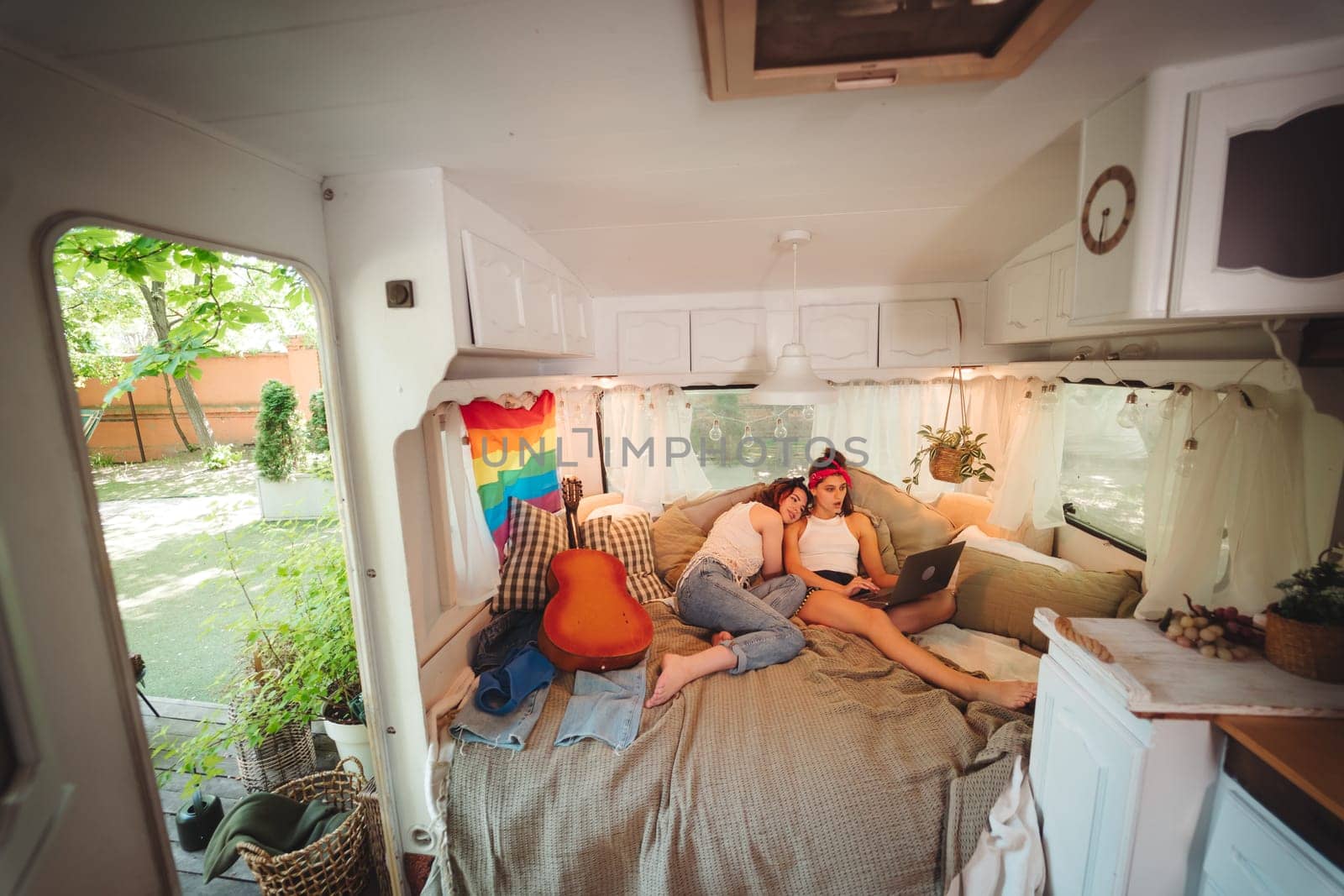 Portrait of a cute lesbian couple. Two girls spend time tenderly together watching movie on laptop in a camper trailer with LGBT flag on the wall. Love and attitude. LGBT concept. High quality photo