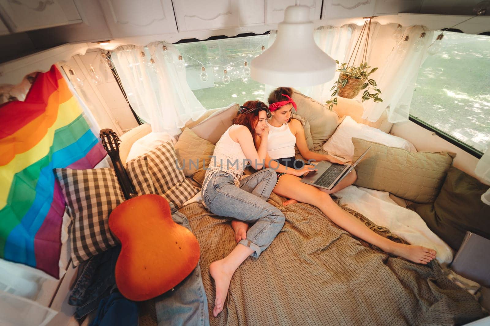 Portrait of a cute lesbian couple. Two girls spend time tenderly together watching movie on laptop in a camper trailer with LGBT flag on the wall. Love and attitude. LGBT concept. High quality photo