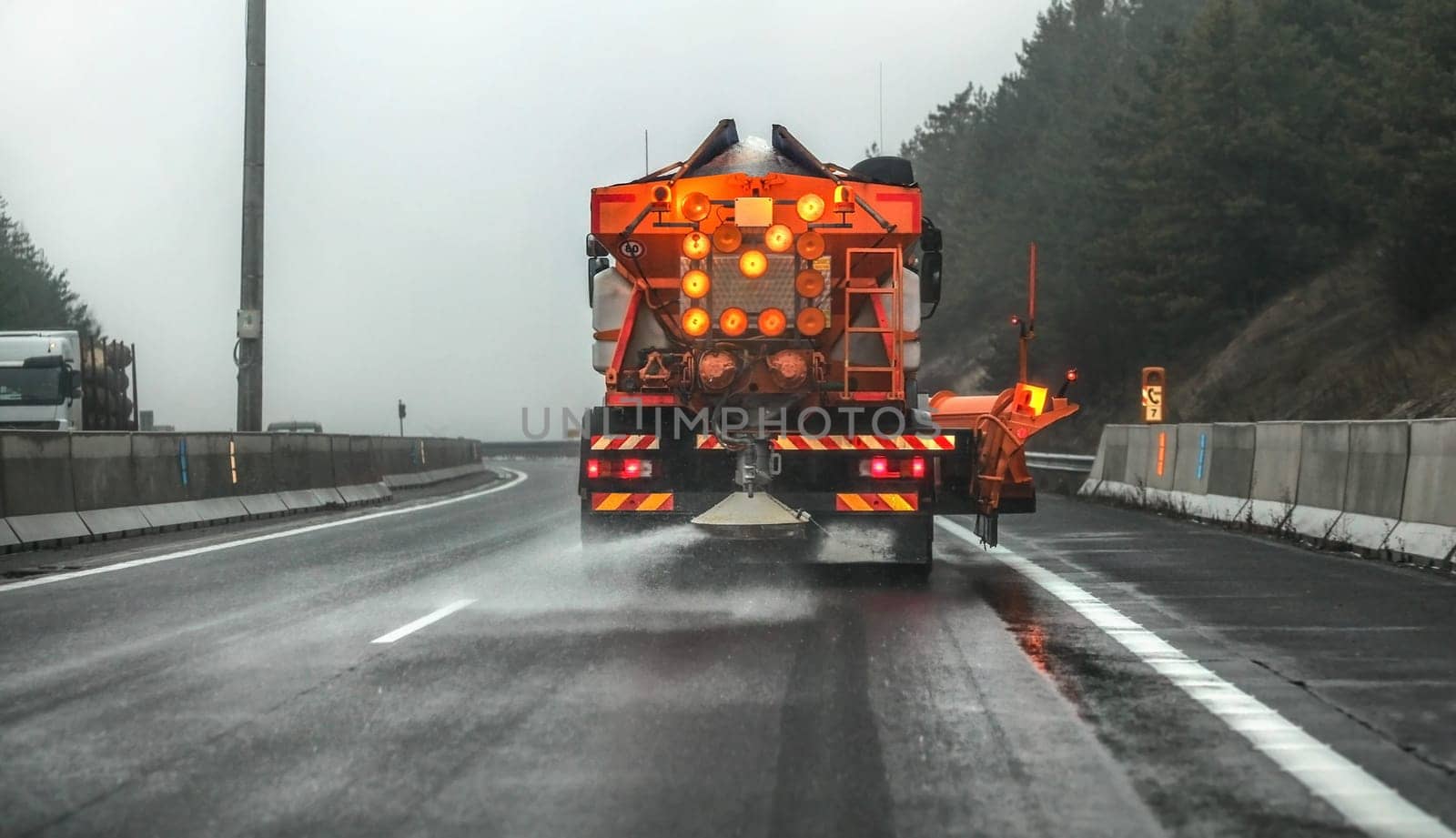 Orange highway maintenance gritter truck spreading de-icing salt, crystals dropping on the ice covered asphalt road during overcast day