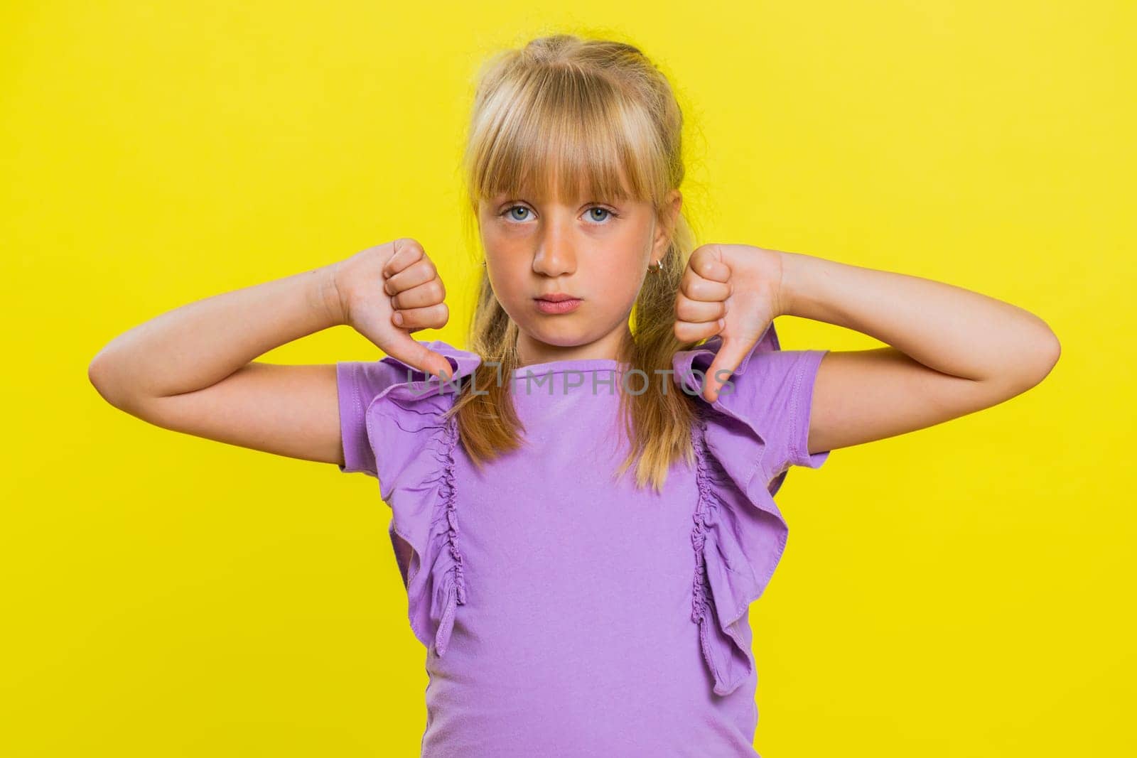Dislike. Upset young preteen child girl kid showing thumbs down sign gesture, expressing discontent, disapproval, dissatisfied bad work indoors. Displeased cute children on yellow studio background