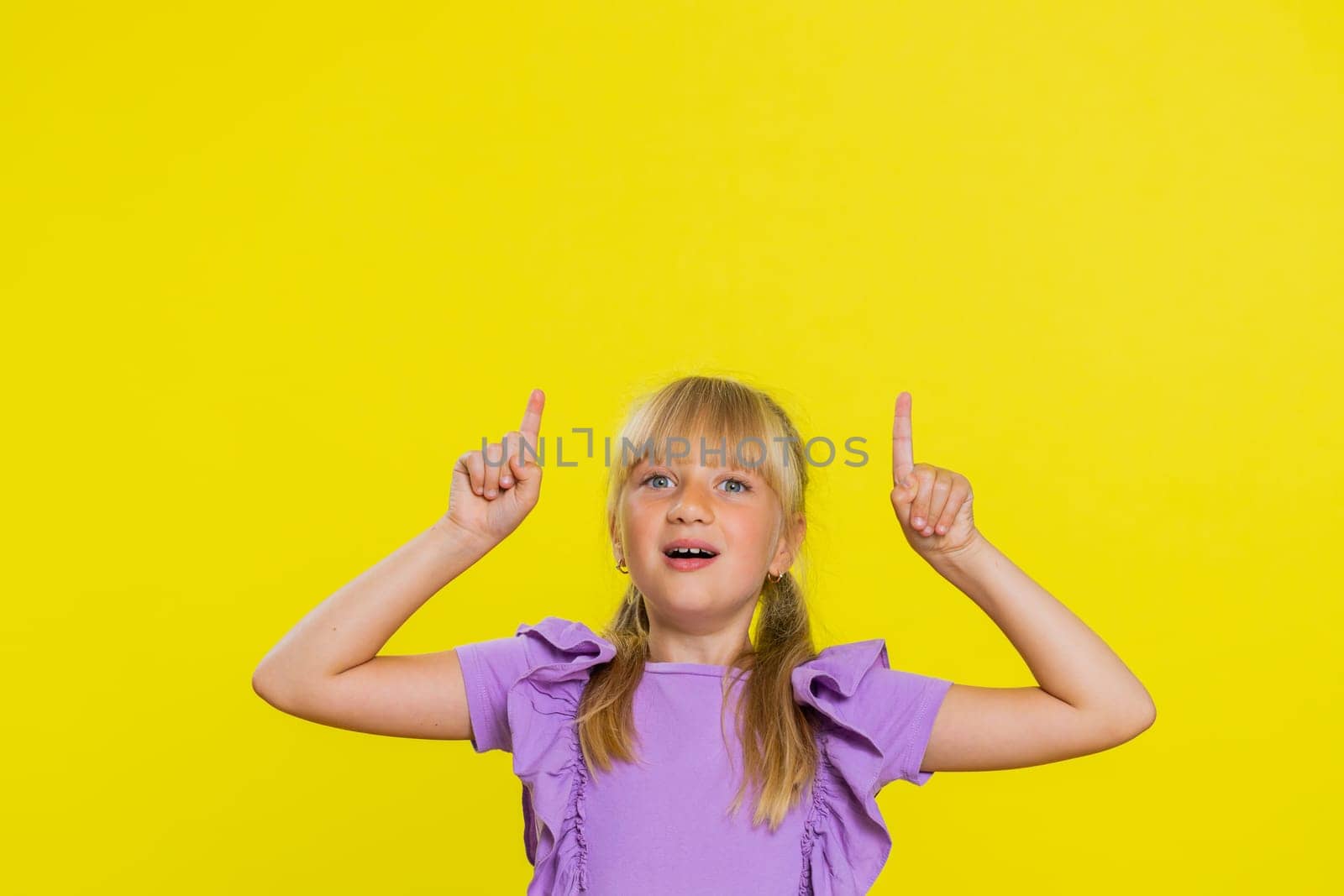 Happy cute girl showing thumbs up pointing empty place up, advertising area for commercial text, copy space for goods promotion banner, copy-space. Young child kid. Studio shot on yellow background