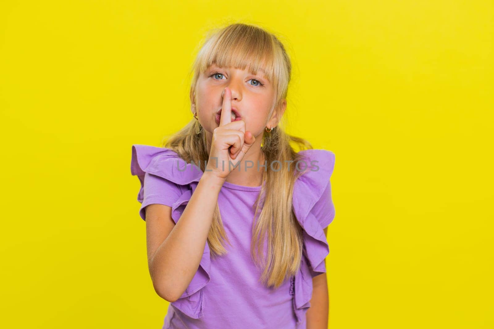Shh be quiet please. Preteen child girl kid presses index finger to lips makes silence hush gesture sign do not tells gossip secret. Little cute children isolated alone on studio yellow background