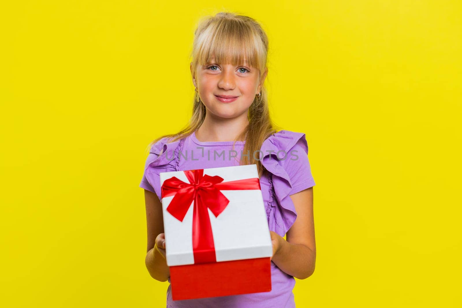 Lovely smiling young preteen child girl kid presenting birthday gift box stretches out hands, offer wrapped present career bonus, celebrating party. Little cute children on studio yellow background