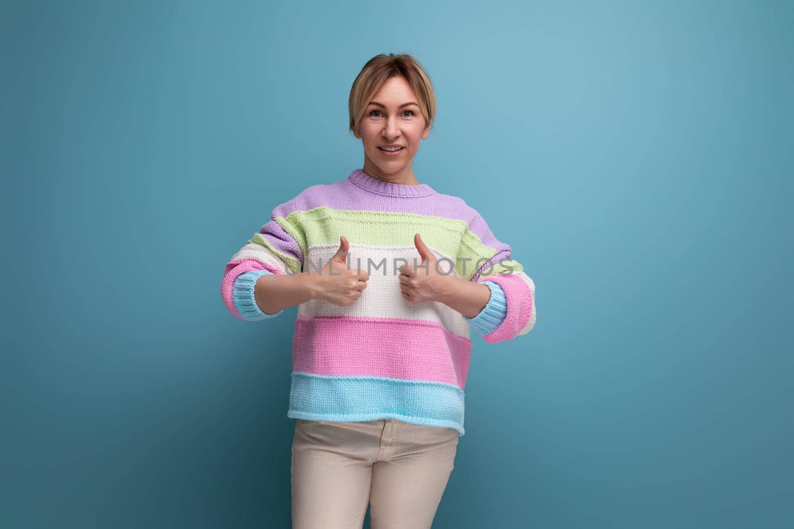 smiling pleasant blond young woman in a striped sweater shows like with her hand on a blue background.