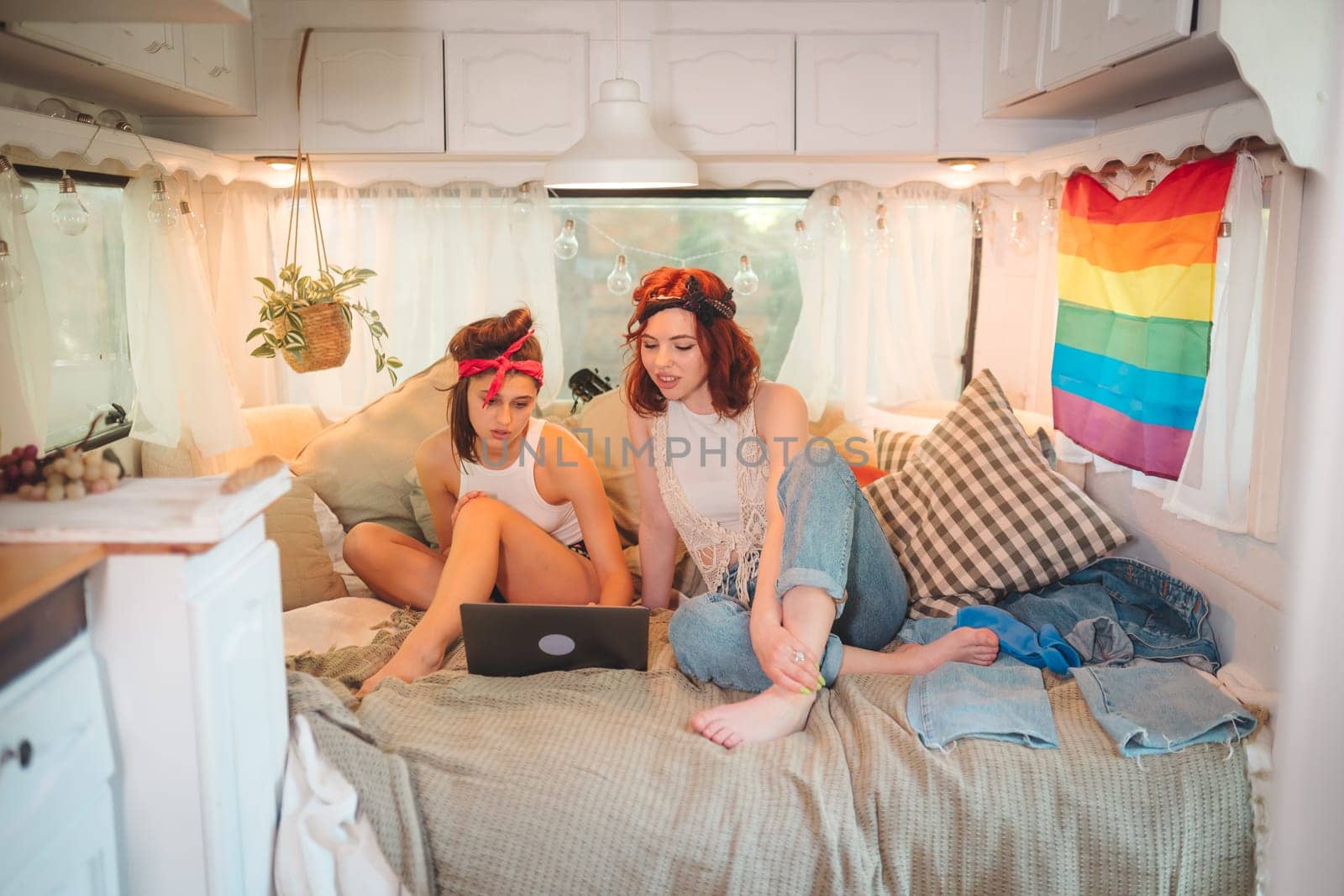 Portrait of a cute lesbian couple. Two girls spend time tenderly together watching movie on laptop in a camper trailer with LGBT flag on the wall. Love and attitude. LGBT concept. High quality photo