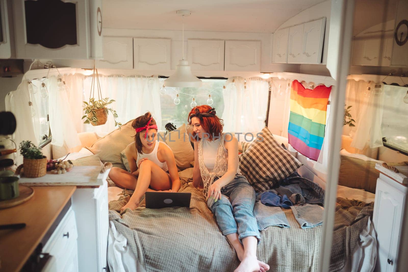 Portrait of a cute lesbian couple. Two girls spend time tenderly together watching movie on laptop in a camper trailer with LGBT flag on the wall. Love and attitude. LGBT concept. High quality photo