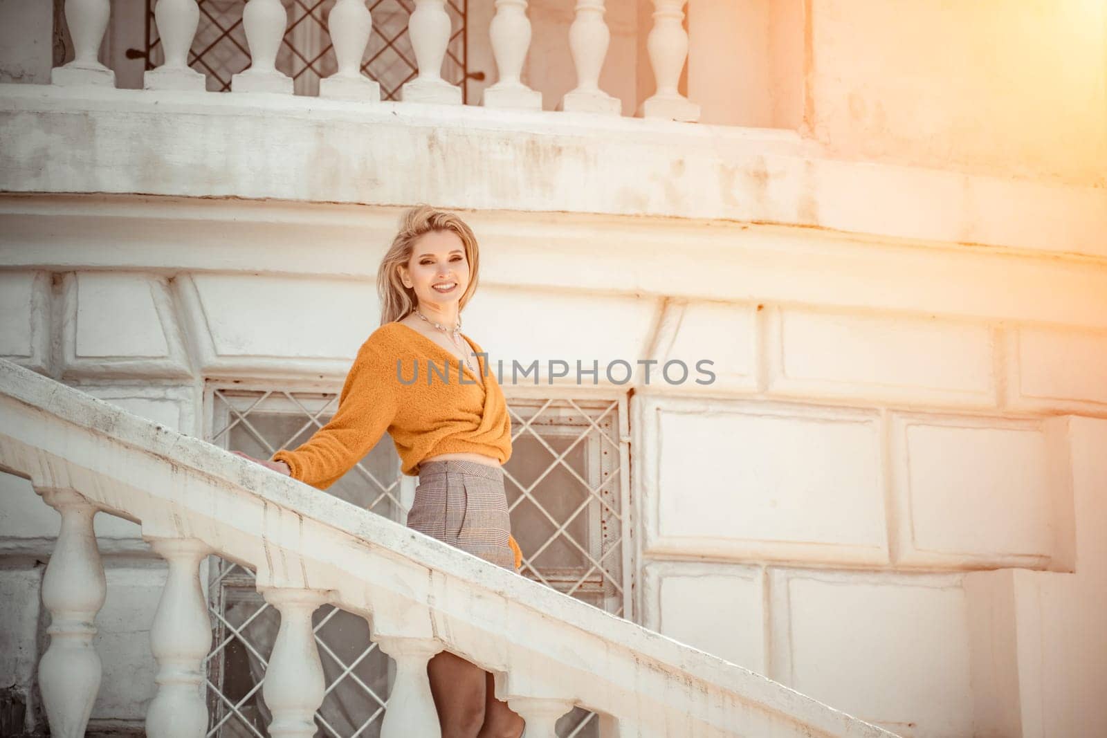 A middle-aged woman looks like a good blonde with curly beautiful hair and makeup on the background of the building. She is wearing a yellow sweater