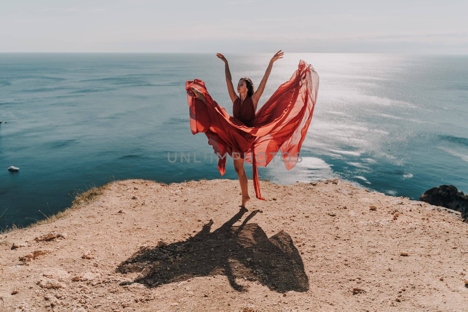 Woman red dress sea. Female dancer posing on a rocky outcrop high above the sea. Girl on the nature on blue sky background. Fashion photo