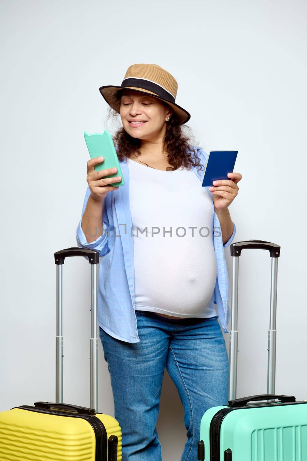 Happy pregnant woman checking her flight on mobile app using her smartphone, going on summer vacations, standing with a boarding pass and suitcases, smiling cutely, isolated on white studio background