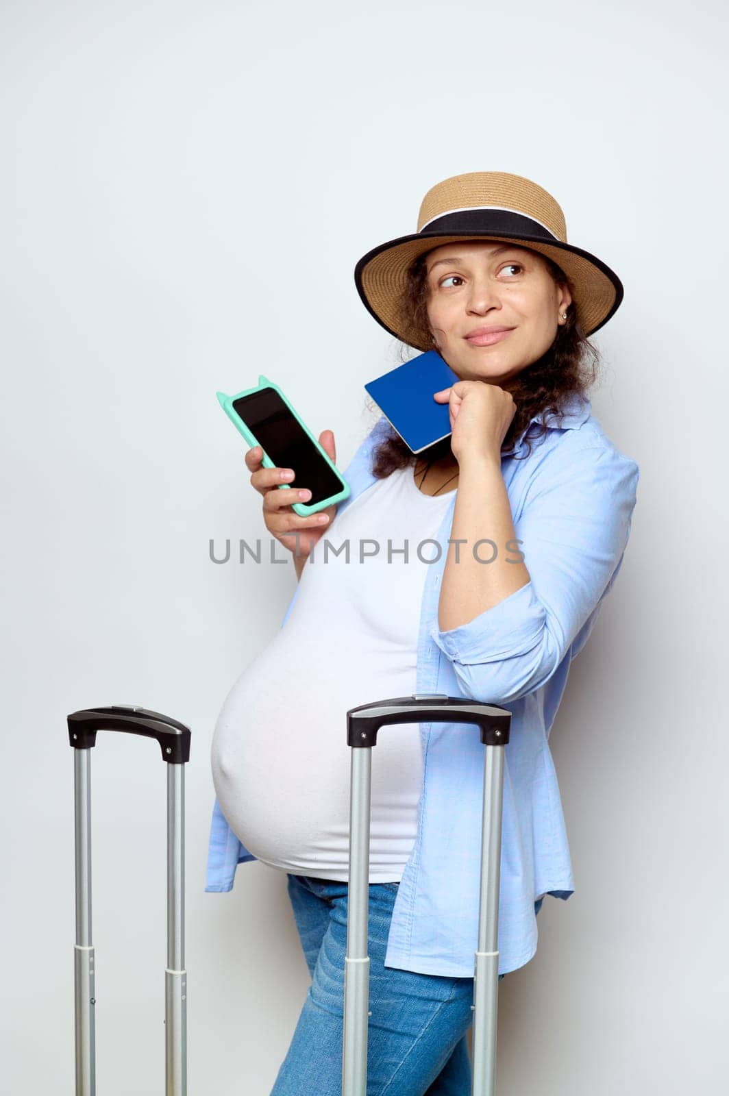 Delightful curly haired pregnant woman with big belly, dreamily looking aside a copy ad space for ads, holding smartphone and passport, isolated white background. Travel by air in late pregnancy time