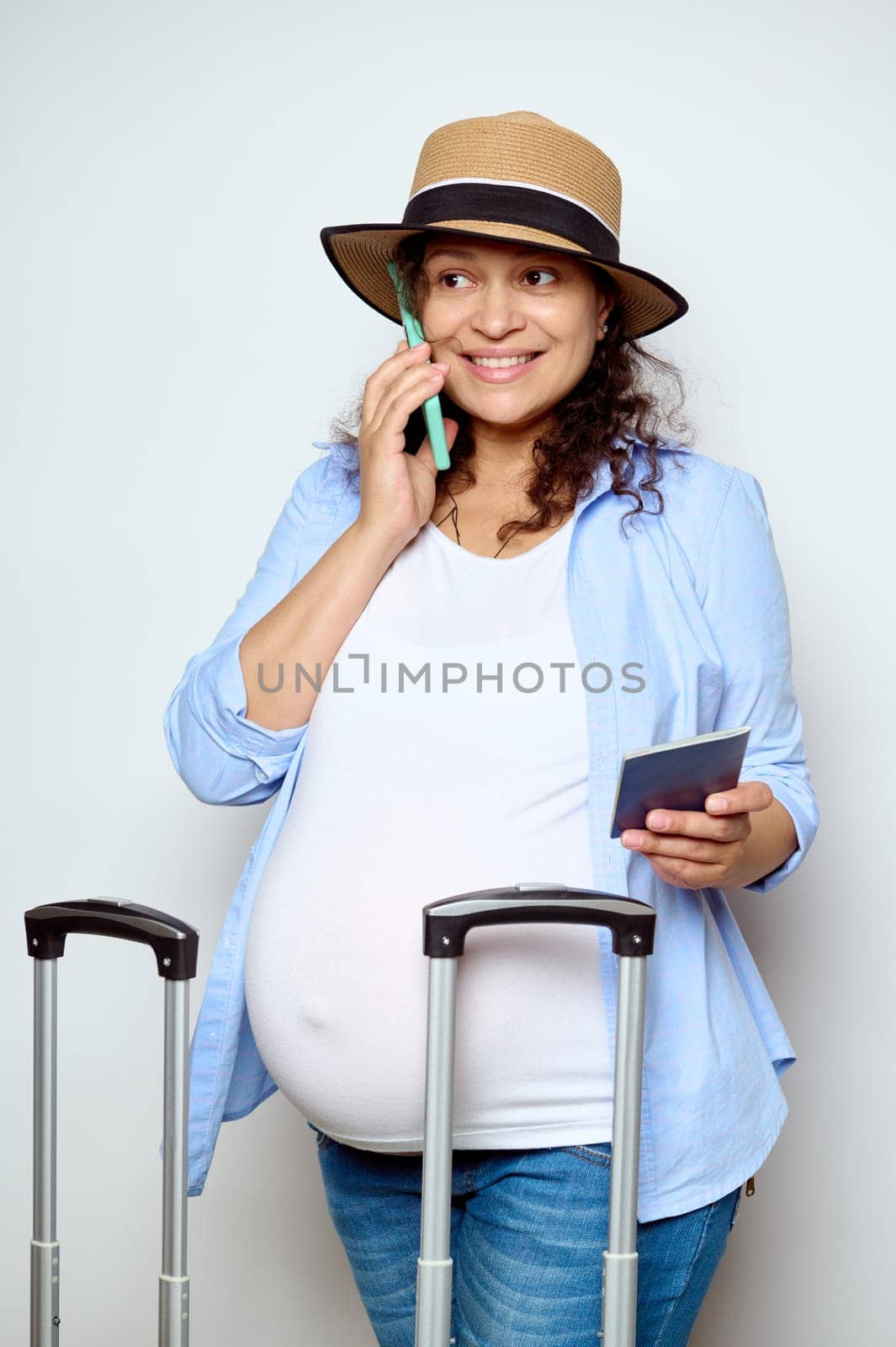 Smiling Latin American pregnant woman holds her passport document, talks on mobile phone, booking hotel, making reservations, standing isolated over white studio background. People. Travel. Pregnancy