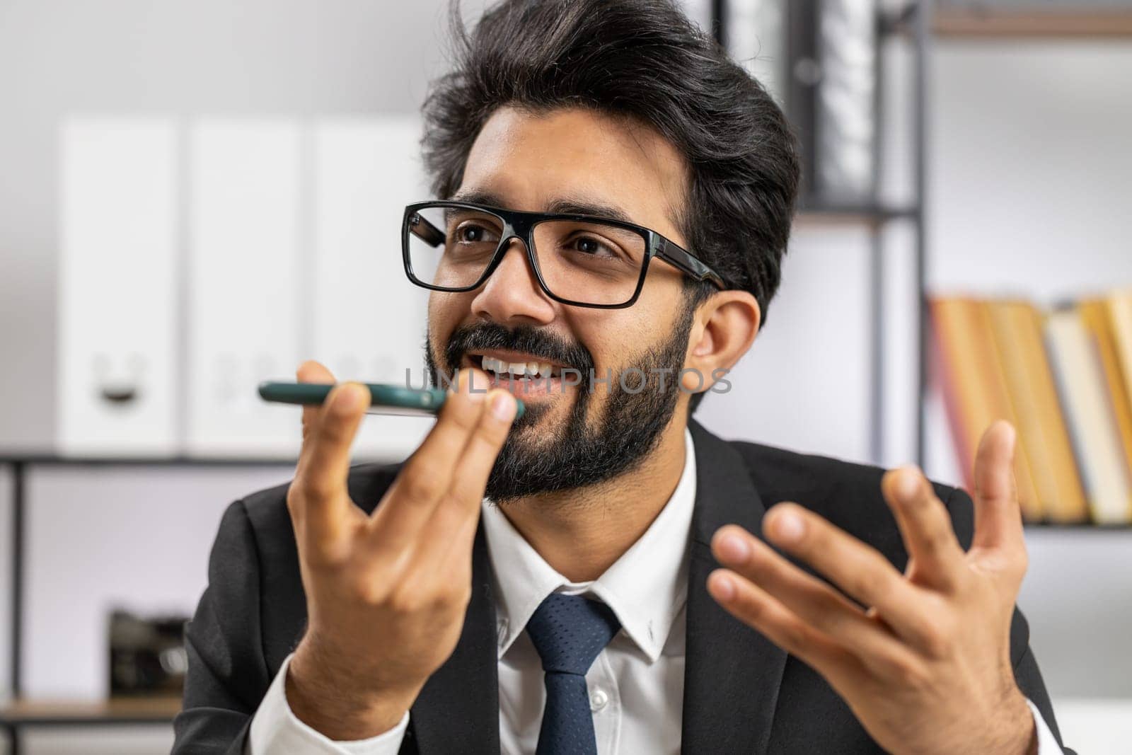 Bearded indian businessman working, having mobile phone loudspeaker talk at office desk with laptop. Manager freelance indian man holding smartphone using messenger chat apps. Employment, occupation