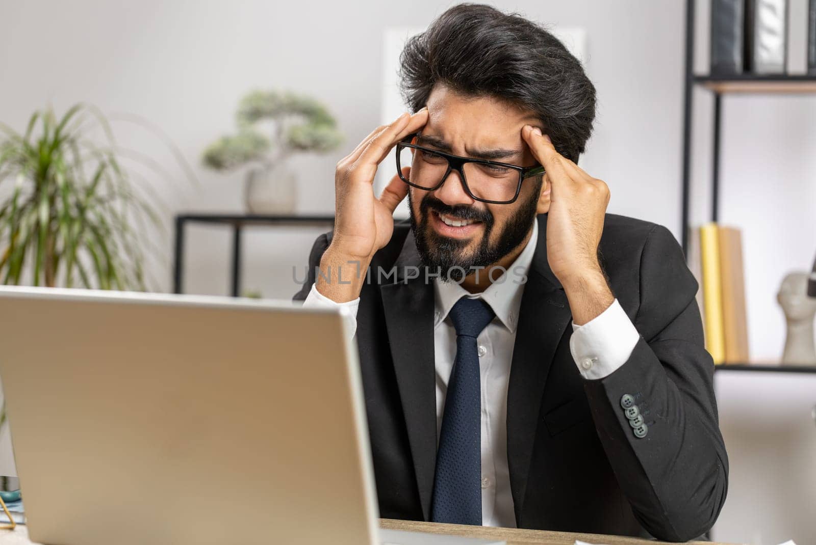 Tired ill indian businessman suffering from headache problem tension and migraine, stress at home office desk workplace. Bearded freelancer broker worker man works on laptop notebook. Overworking