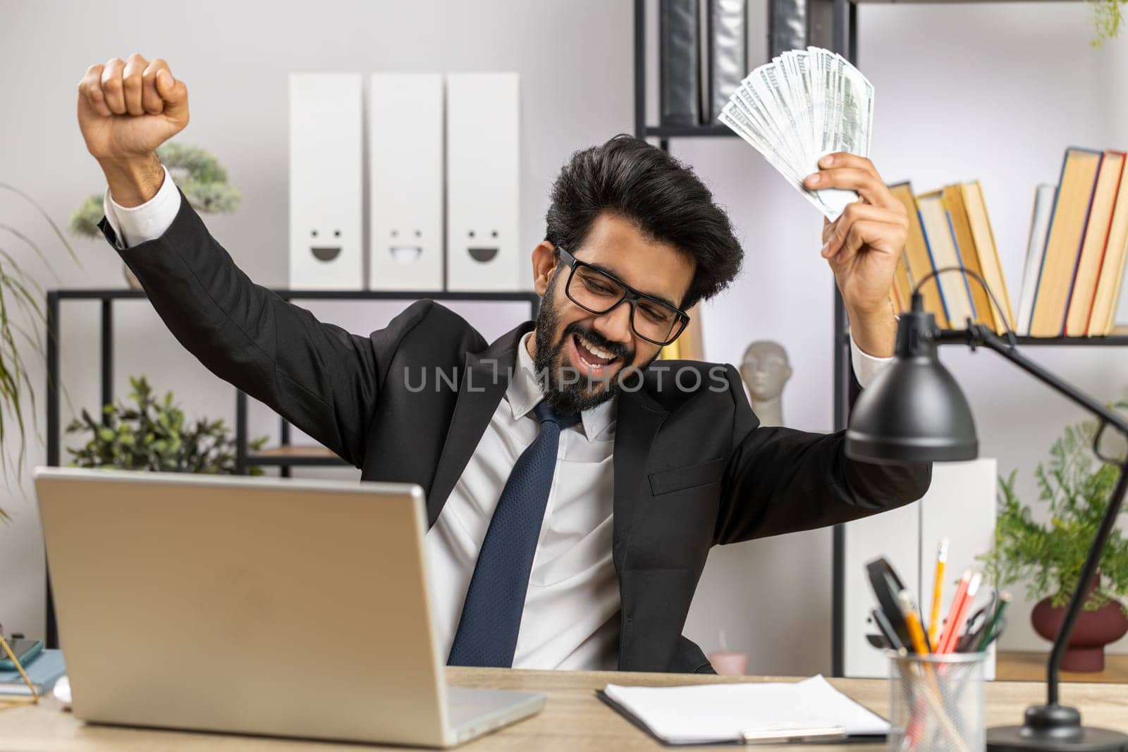 Indian businessman holding fan of cash money dollar banknotes celebrate, dance, success career, lottery jackpot game winner, big income, wealth at home office workplace desk. Remote distant working