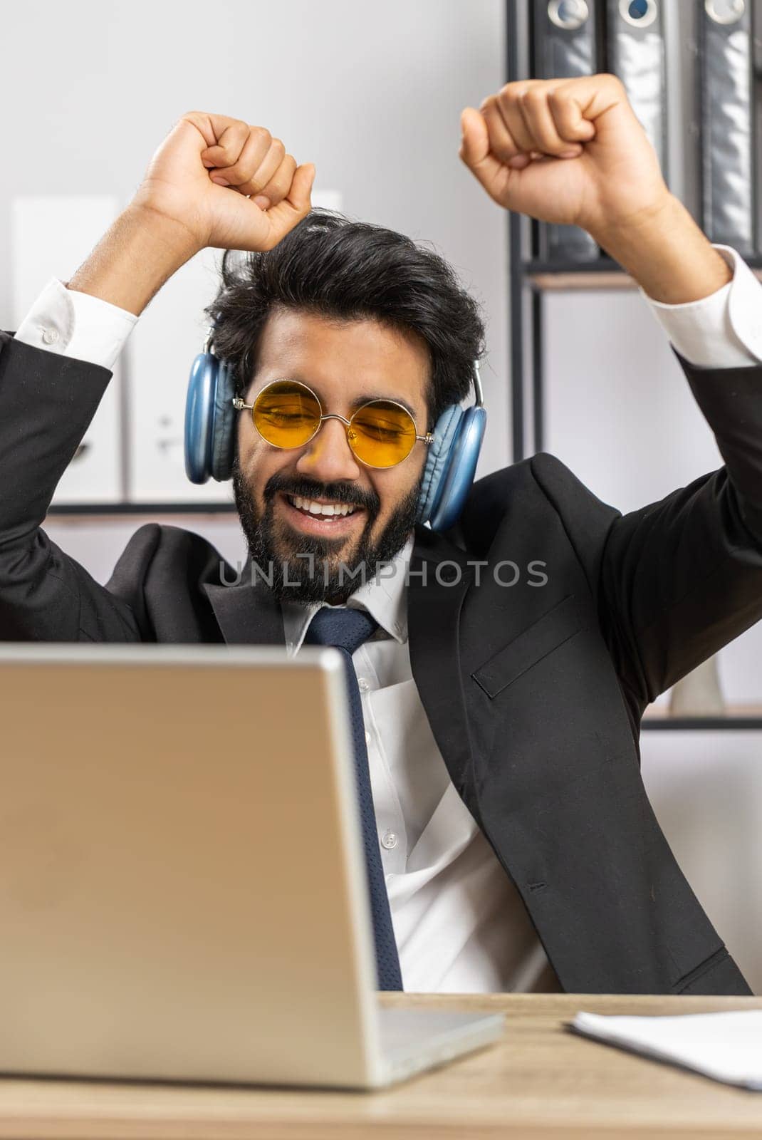 Happy relaxed overjoyed indian businessman working on laptop computer at home office wearing headphones listening favorite energetic disco music and dancing. Freelancer man relaxing, taking a break