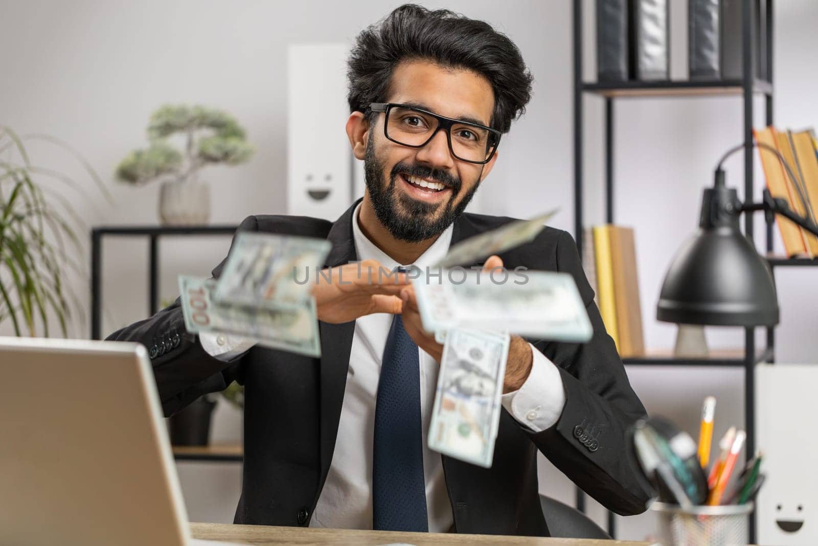 Cheerful rich young indian businessman working on laptop and wasting throwing money to camera more tips earnings big profit success win lottery shopping. Happy freelancer arab man at home office desk